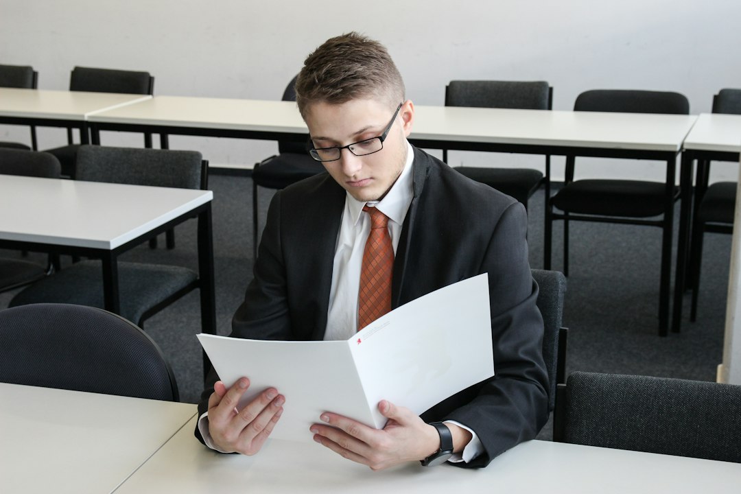 Professor in a dark suit and glasses reviewing course documents at an empty classroom desk