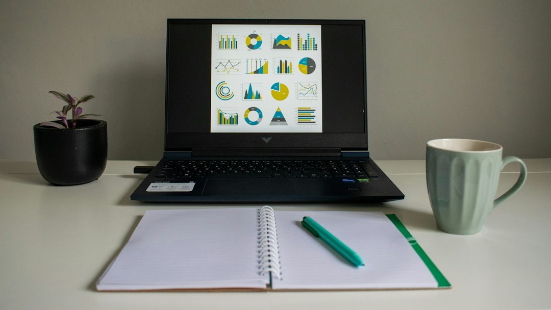 A laptop displaying colorful data charts and dashboards next to an open notebook and coffee mug on a desk