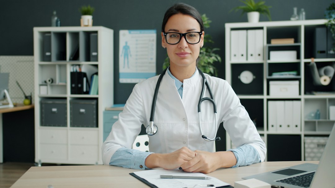 A medical professional seated at an office desk with binders and documents behind them